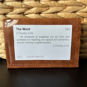 May include: A brown leather card holder with a clear plastic window, containing a white card with the text "The Word" and a Bible verse. The card holder is on a woven background.
