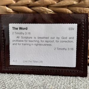 Puede incluir: Una cartera de cuero marrón oscuro con una ventana de plástico transparente, que muestra una tarjeta blanca con texto de 2 Timoteo 3:16. El texto dice "The Word" y "Live the New Life". La cartera está sobre una superficie texturizada.