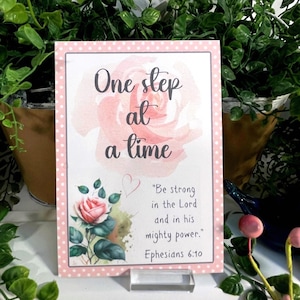 May include: A decorative print with a pink floral design and the text "One step at a time." The print also includes the quote "Be strong in the Lord and in his mighty power." from Ephesians 6:10.