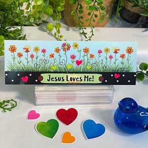 K&ouml;nnte beinhalten: Eine rechteckige Karte mit hellblauem Himmel, gr&uuml;nem Gras und bunten Blumen sowie dem Text "Jesus Loves Me!" auf schwarzem Grund mit wei&szlig;en Punkten. Unter der Karte liegen f&uuml;nf kleine herzf&ouml;rmige Ausschnitte und ein blauer Glasvogel.