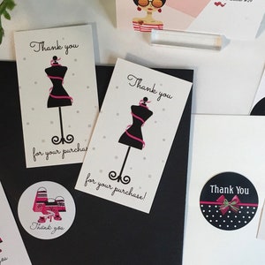 May include: Thank you cards with a black and white illustration of a dress form with a pink ribbon. The text "Thank you for your purchase!" is printed on the cards. The cards are on a black surface.