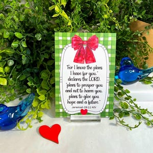 K&ouml;nnte beinhalten: Eine rechteckige Karte mit gr&uuml;n-wei&szlig; kariertem Hintergrund, einer roten Schleife oben und einem kleinen roten Herz unten. Der Text lautet: For I know the plans I have for you, declares the LORD, plans to prosper you and not to harm you, plans to give you hope and a future. Jeremiah 29:11 NIV.
