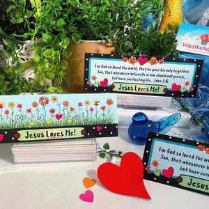 K&ouml;nnte beinhalten: Drei rechteckige Karten mit hellblauem Hintergrund, bunten Blumen, gr&uuml;nem Gras und roten Herzen. Der Text lautet "Jesus Loves Me!" und enth&auml;lt den Bibelvers Johannes 3,16. Ein gro&szlig;es rotes Herz ist im Vordergrund platziert.