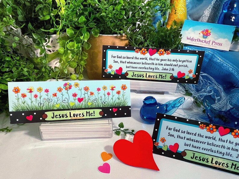 K&ouml;nnte beinhalten: Drei rechteckige Karten mit hellblauem Hintergrund, bunten Blumen, gr&uuml;nem Gras und roten Herzen. Der Text lautet "Jesus Loves Me!" und enth&auml;lt den Bibelvers Johannes 3,16. Ein gro&szlig;es rotes Herz ist im Vordergrund platziert.