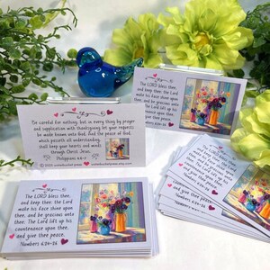 Pode incluir: Um conjunto de pequenos cart&otilde;es com obras de arte florais e vers&iacute;culos b&iacute;blicos. Os cart&otilde;es apresentam uma pintura de flores em vasos e texto da B&iacute;blia. Uma estatueta de p&aacute;ssaro de vidro azul est&aacute; colocada perto, com folhagem verde e flores amarelas no fundo.