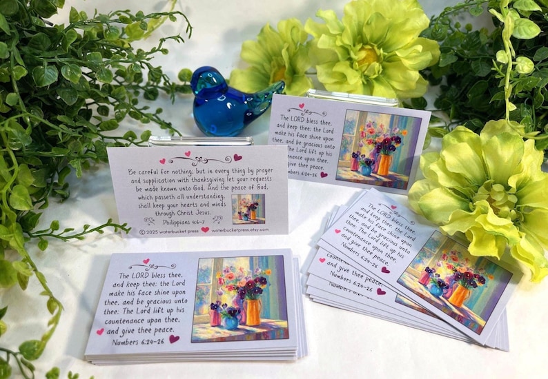 Pode incluir: Um conjunto de pequenos cart&otilde;es com obras de arte florais e vers&iacute;culos b&iacute;blicos. Os cart&otilde;es apresentam uma pintura de flores em vasos e texto da B&iacute;blia. Uma estatueta de p&aacute;ssaro de vidro azul est&aacute; colocada perto, com folhagem verde e flores amarelas no fundo.