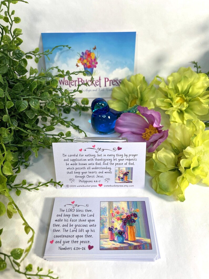 Pode incluir: Uma figura de p&aacute;ssaro de vidro azul est&aacute; perto de arranjos florais e cart&otilde;es com vers&iacute;culos b&iacute;blicos. Os cart&otilde;es apresentam pinturas florais coloridas e texto de Filipenses 4:6-7 e N&uacute;meros 6:24-26. O fundo inclui um cart&atilde;o com o texto "WaterBucket Press."