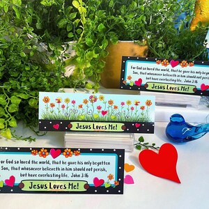 K&ouml;nnte beinhalten: Drei rechteckige Lesezeichen mit schwarzem Rand und wei&szlig;en Punkten, bunten Blumenillustrationen und roten Herzakzenten. Zwei Lesezeichen zeigen den Text 'For God so loved the world' und 'Jesus Loves Me!', eines zeigt lediglich Blumen.