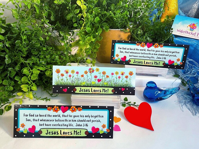 K&ouml;nnte beinhalten: Drei rechteckige Lesezeichen mit schwarzem Rand und wei&szlig;en Punkten, bunten Blumenillustrationen und roten Herzakzenten. Zwei Lesezeichen zeigen den Text 'For God so loved the world' und 'Jesus Loves Me!', eines zeigt lediglich Blumen.