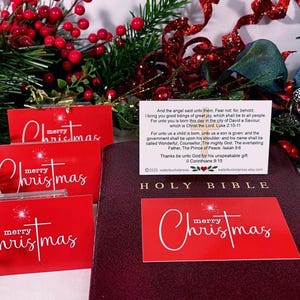Peut inclure: Cartes de No&euml;l rouges avec l'inscription "merry Christmas" en blanc. Une Sainte Bible bordeaux et des d&eacute;corations festives avec des baies rouges et des rubans sont en arri&egrave;re-plan. Une petite carte avec du texte est &eacute;galement pr&eacute;sente.