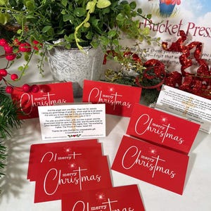 Peut inclure: Cartes de No&euml;l rouges avec l'inscription "merry Christmas" en &eacute;criture blanche. Les cartes sont dispos&eacute;es avec de la verdure et des baies festives. Une petite plante dans un pot argent&eacute; est &eacute;galement visible.