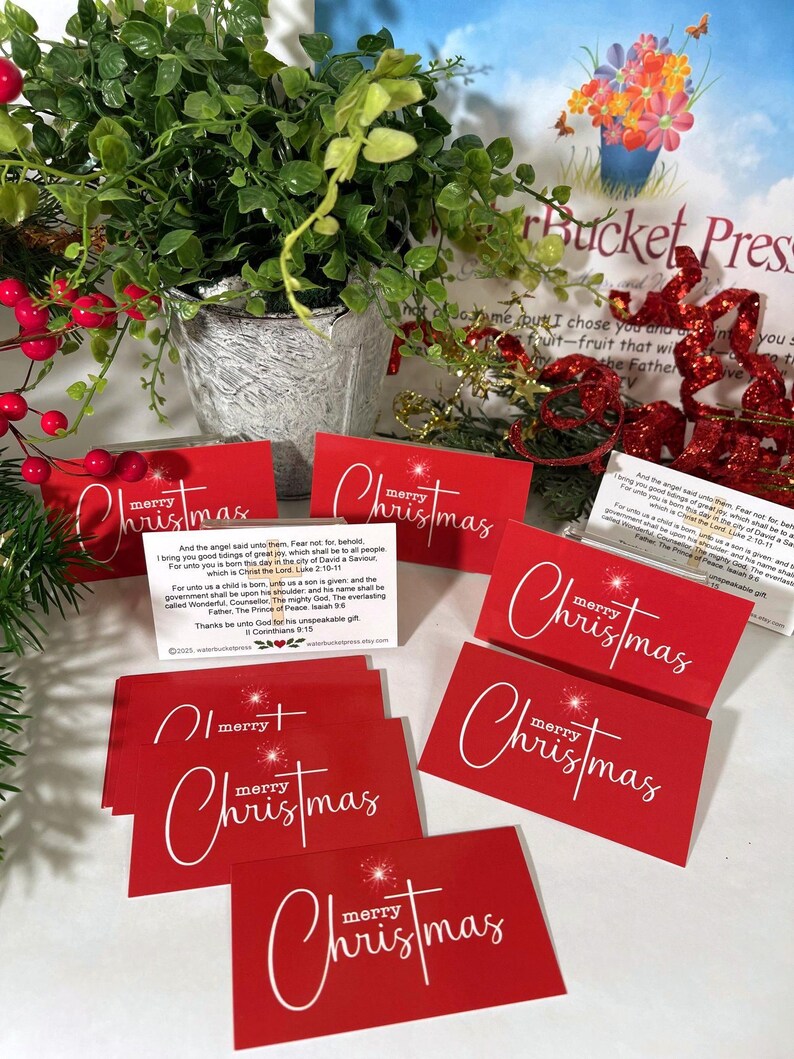 Peut inclure: Cartes de No&euml;l rouges avec l'inscription "merry Christmas" en &eacute;criture blanche. Les cartes sont dispos&eacute;es avec de la verdure et des baies festives. Une petite plante dans un pot argent&eacute; est &eacute;galement visible.