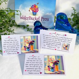 Pode incluir: A imagem apresenta tr&ecirc;s cart&otilde;es com arte floral e vers&iacute;culos b&iacute;blicos, juntamente com duas estatuetas de p&aacute;ssaros de vidro azul e um letreiro que diz "WaterBucket Press". Os cart&otilde;es t&ecirc;m uma pintura em aquarela de flores em vasos.