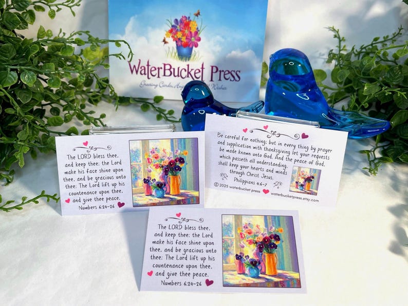 Pode incluir: A imagem apresenta tr&ecirc;s cart&otilde;es com arte floral e vers&iacute;culos b&iacute;blicos, juntamente com duas estatuetas de p&aacute;ssaros de vidro azul e um letreiro que diz "WaterBucket Press". Os cart&otilde;es t&ecirc;m uma pintura em aquarela de flores em vasos.