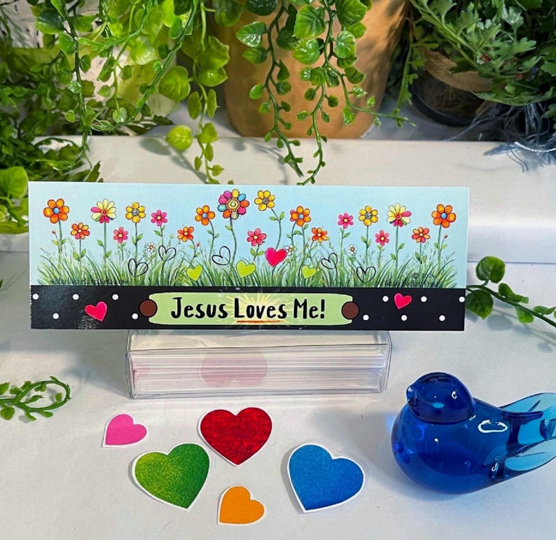K&ouml;nnte beinhalten: Eine rechteckige Karte mit hellblauem Himmel, gr&uuml;nem Gras und bunten Blumen sowie dem Text "Jesus Loves Me!" auf schwarzem Grund mit wei&szlig;en Punkten. Unter der Karte liegen f&uuml;nf kleine herzf&ouml;rmige Ausschnitte und ein blauer Glasvogel.