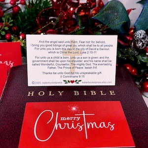 Peut inclure: Une Sainte Bible bordeaux avec des lettres dor&eacute;es, une carte de No&euml;l rouge et une carte blanche avec du texte. La carte de No&euml;l porte l'inscription "Joyeux No&euml;l" en &eacute;criture blanche. La carte blanche contient du texte de Luc 2:10-11 et d'Isa&iuml;e 9:6.