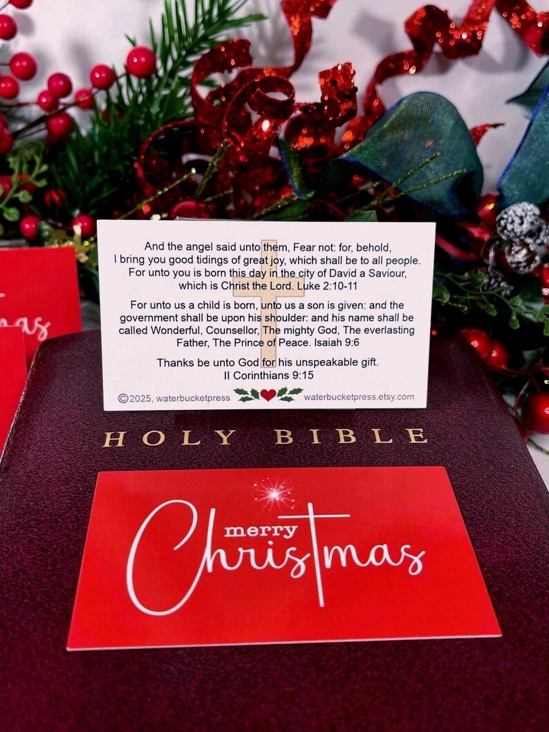 Peut inclure: Une Sainte Bible bordeaux avec des lettres dor&eacute;es, une carte de No&euml;l rouge et une carte blanche avec du texte. La carte de No&euml;l porte l'inscription "Joyeux No&euml;l" en &eacute;criture blanche. La carte blanche contient du texte de Luc 2:10-11 et d'Isa&iuml;e 9:6.