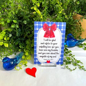 K&ouml;nnte beinhalten: Eine rechteckige Karte mit blau-wei&szlig;em Vichy-Muster, verziert mit einer roten Schleife oben und einem kleinen roten Herz unten. Der Text lautet: I will be glad and rejoice in your unfailing love, for you have seen my troubles, and you care about the anguish of my soul. Psalm 31:7 NLT.