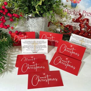 Peut inclure: Des cartes de No&euml;l rouges avec l'inscription "merry Christmas" en blanc. Certaines cartes pr&eacute;sentent une &eacute;toile. Les cartes sont dispos&eacute;es avec de la verdure et des baies rouges, cr&eacute;ant une ambiance festive.