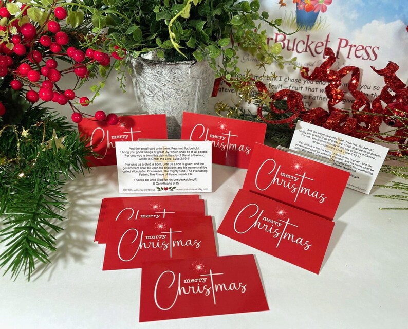 Peut inclure: Des cartes de No&euml;l rouges avec l'inscription "merry Christmas" en blanc. Certaines cartes pr&eacute;sentent une &eacute;toile. Les cartes sont dispos&eacute;es avec de la verdure et des baies rouges, cr&eacute;ant une ambiance festive.