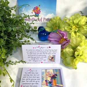 Pode incluir: Uma figura de p&aacute;ssaro de vidro azul est&aacute; perto de arranjos florais e cart&otilde;es com vers&iacute;culos b&iacute;blicos. Os cart&otilde;es apresentam pinturas florais coloridas e texto de Filipenses 4:6-7 e N&uacute;meros 6:24-26. O fundo inclui um cart&atilde;o com o texto "WaterBucket Press."
