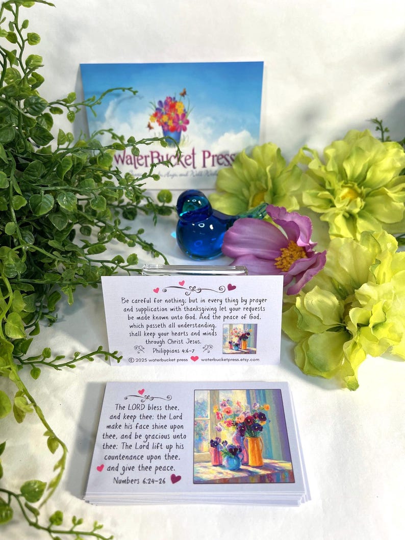 Pode incluir: Uma figura de p&aacute;ssaro de vidro azul est&aacute; perto de arranjos florais e cart&otilde;es com vers&iacute;culos b&iacute;blicos. Os cart&otilde;es apresentam pinturas florais coloridas e texto de Filipenses 4:6-7 e N&uacute;meros 6:24-26. O fundo inclui um cart&atilde;o com o texto "WaterBucket Press."