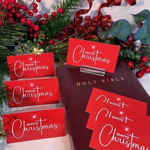 Peut inclure: Cartes de No&euml;l rouges avec une &eacute;criture blanche et un motif d'&eacute;clat d'&eacute;toile, pr&eacute;sent&eacute;es pr&egrave;s d'une Sainte Bible bordeaux. De la verdure festive et des baies rouges composent le d&eacute;cor. Les cartes sont dans des pr&eacute;sentoirs en acrylique transparent.