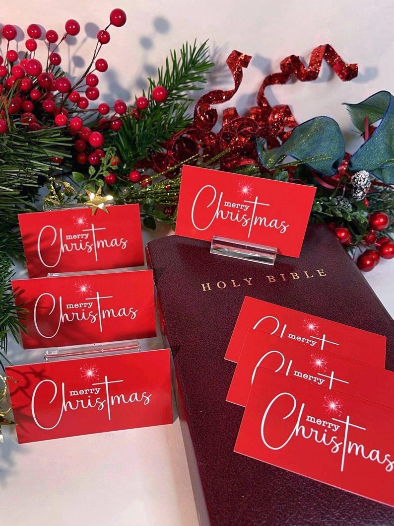 Peut inclure: Cartes de No&euml;l rouges avec une &eacute;criture blanche et un motif d'&eacute;clat d'&eacute;toile, pr&eacute;sent&eacute;es pr&egrave;s d'une Sainte Bible bordeaux. De la verdure festive et des baies rouges composent le d&eacute;cor. Les cartes sont dans des pr&eacute;sentoirs en acrylique transparent.