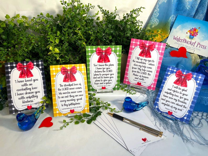 K&ouml;nnte beinhalten: F&uuml;nf rechteckige Karten mit Vichy-Karo-R&auml;ndern in Schwarz, Gelb, Gr&uuml;n, Pink und Blau, jede mit einer roten Schleifenillustration, einem wei&szlig;en Mittelfeld mit gedrucktem religi&ouml;sem Text und einem kleinen roten Herzsymbol unten.