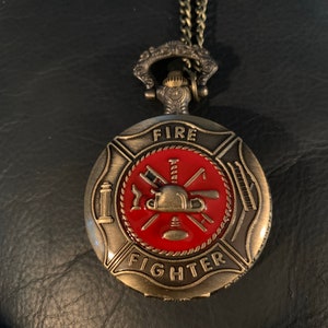 May include: A gold-toned pocket watch with a chain. The watch face features a red enamel firefighter emblem with a crossed axe and a helmet. The words "FIRE FIGHTER" are inscribed around the emblem.