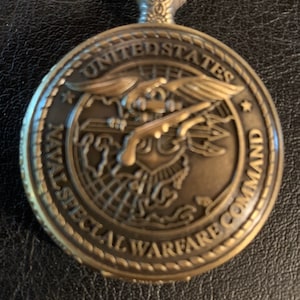 May include: A gold-toned pocket watch with a detailed design featuring the United States Naval Special Warfare Command emblem. The emblem depicts a stylized eagle with wings spread, holding a trident and a rifle, with a globe in the background.