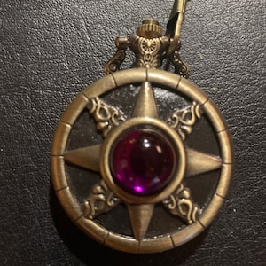May include: A gold-toned pocket watch with a purple gem in the center. The watch face features a star design with ornate details around the edge.