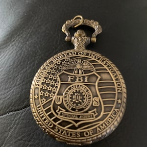 May include: A gold-toned pocket watch with a chain attached. The watch face features the FBI seal with the words "Federal Bureau of Investigation" and "United States of America" around the edge.