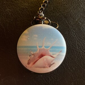 May include: A silver pocket watch with a chain. The watch face has a picture of a seashell on a beach.