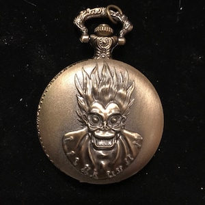 May include: A gold-toned pocket watch with a detailed image of a character with a skull-like face and spiky hair. The watch has a decorative chain attached to the top.