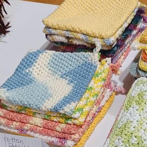 May include: A stack of colorful crocheted cotton pot holders. The pot holders are in various colors, including blue, yellow, green, pink, and white. The sign reads "Cotton Potholders $10.00".