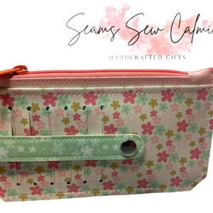May include: A pink and green zippered pouch with a floral print and a strap with a button closure. The pouch has multiple slots for needles or other small items. The text "Seams Sew Calming" and "HANDCRAFTED GIFTS" is visible on the pouch.