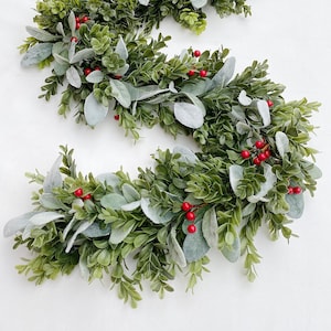 Green dark boxwood style faux greenery garland and lambs ear garland is laying on a white backdrop in a kitchen. Garland is full of both pieces of greenery and has red Christmas holiday style berries distributed throughout.