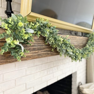 Green eucalyptus and lambs ear garland is laying on a brown wood mantle over a fireplace. Garland is full of both pieces of greenery.