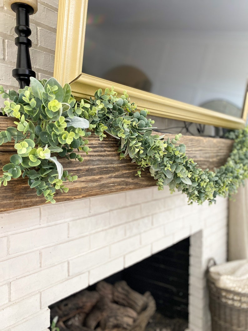 Green eucalyptus and lambs ear garland is laying on a brown wood mantle over a fireplace. Garland is full of both pieces of greenery.