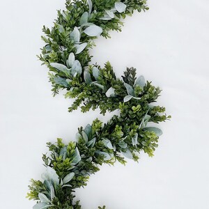 Green dark boxwood style faux greenery garland and lambs ear garland is laying on a white backdrop in a kitchen. Garland is full of both pieces of greenery