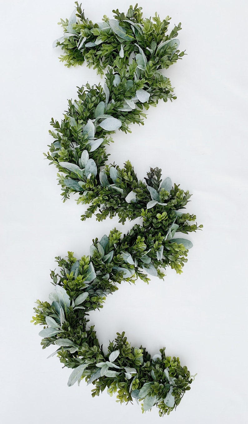 Green dark boxwood style faux greenery garland and lambs ear garland is laying on a white backdrop in a kitchen. Garland is full of both pieces of greenery