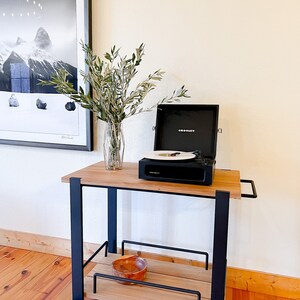 May include: A black record player on a wooden cart with a vase of green branches. The record player is open and has a record on the turntable. The cart has a black metal frame and wooden shelves.