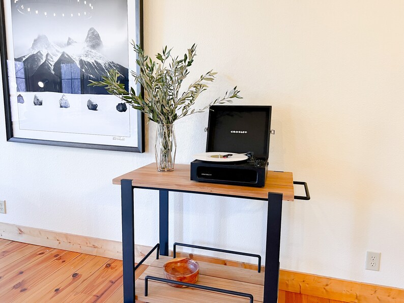 May include: A black record player on a wooden cart with a vase of green branches. The record player is open and has a record on the turntable. The cart has a black metal frame and wooden shelves.