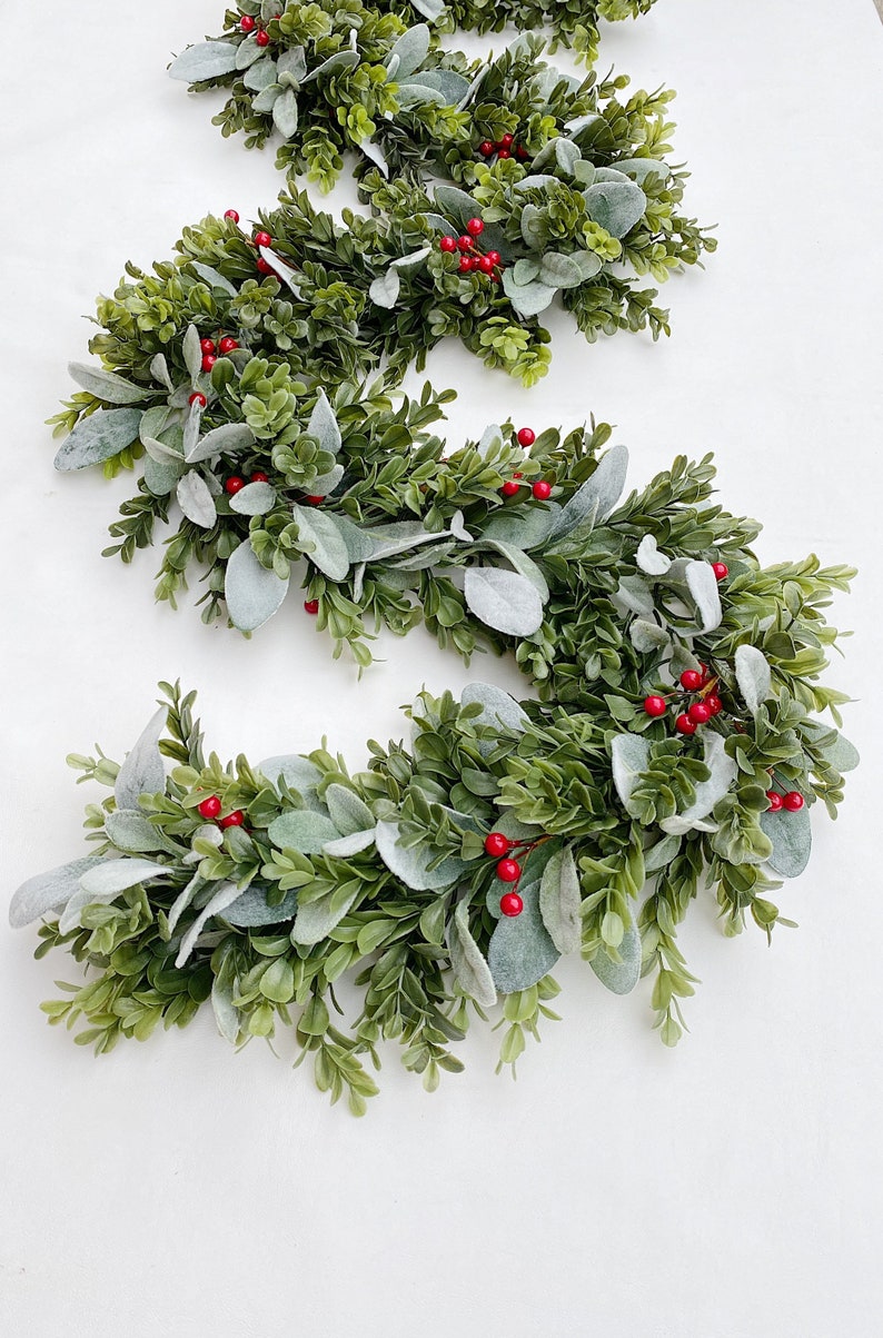 Green dark boxwood style faux greenery garland and lambs ear garland is laying on a white backdrop in a kitchen. Garland is full of both pieces of greenery and has red Christmas holiday style berries distributed throughout.