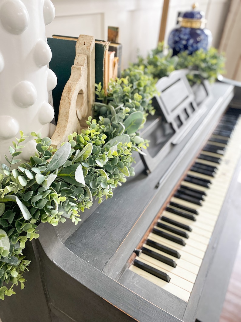 Green eucalyptus and lambs ear garland is laying on the back of a black piano being used as piano decoration. Garland is full of both pieces of greenery.