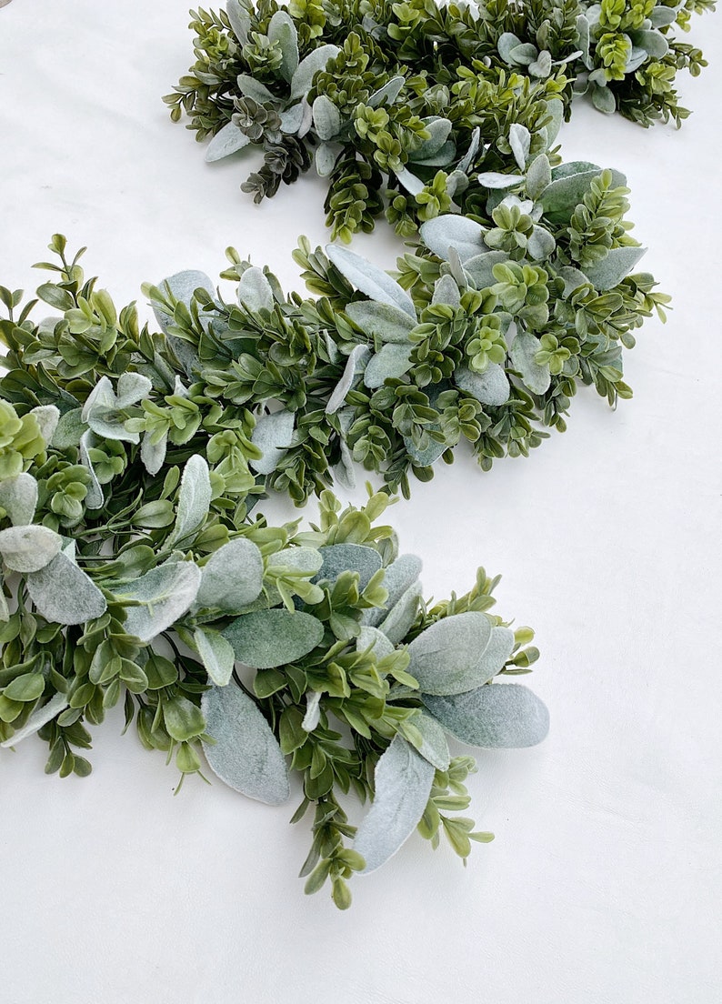 Green dark boxwood style faux greenery garland and lambs ear garland is laying on a white backdrop in a kitchen. Garland is full of both pieces of greenery