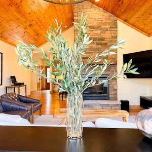 May include: A living room interior with a stone fireplace, a large flat screen TV, and a wooden coffee table. A vase with green branches sits on the table, and a decorative ceramic pot is on the floor.