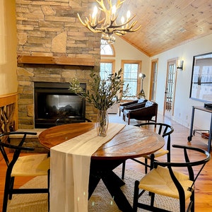 May include: A round wooden dining table with a white runner and four black chairs with woven seats. The table is set in a room with a stone fireplace and a chandelier made of antlers.