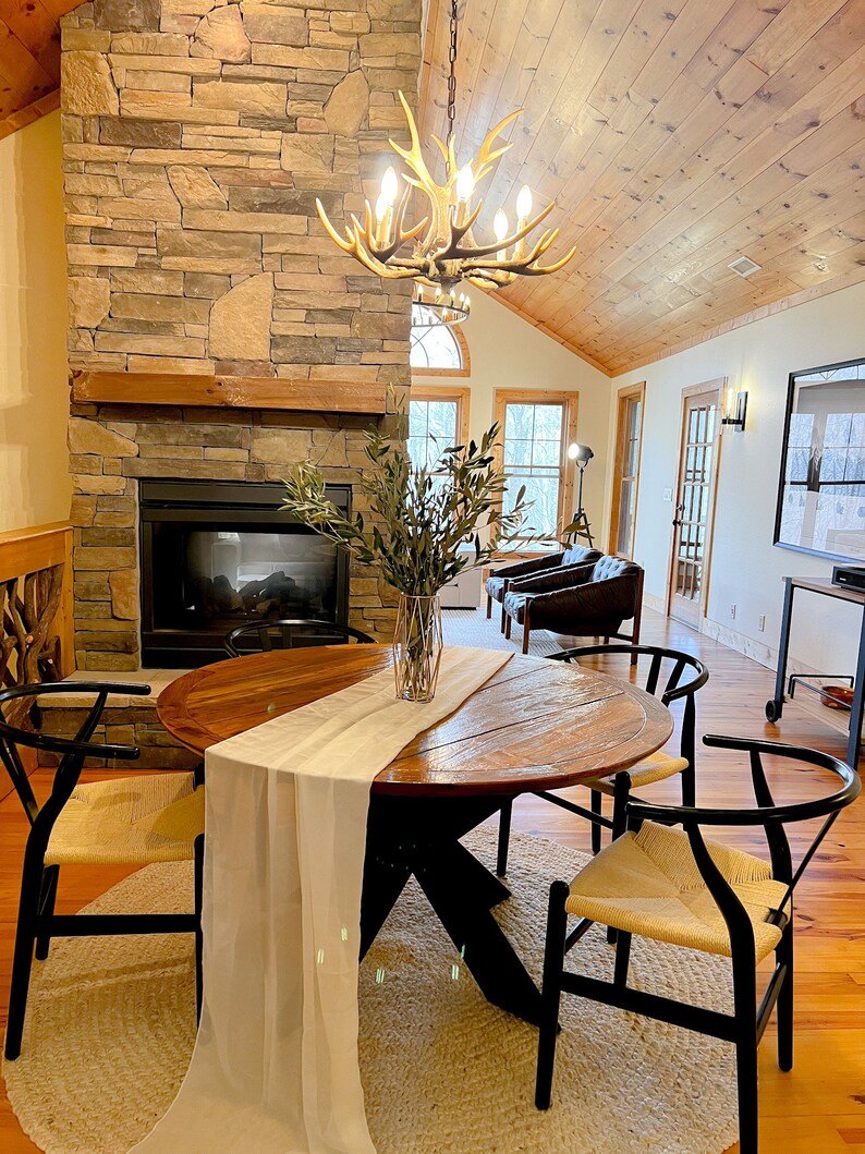 May include: A round wooden dining table with a white runner and four black chairs with woven seats. The table is set in a room with a stone fireplace and a chandelier made of antlers.